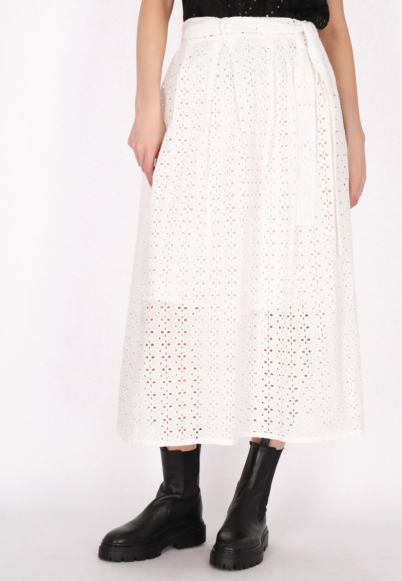 Jupe midi blanche en tissu brodé avec des motifs découpés. Elle présente une ceinture à nouer à la taille et est associée à des bottes noires à la cheville.