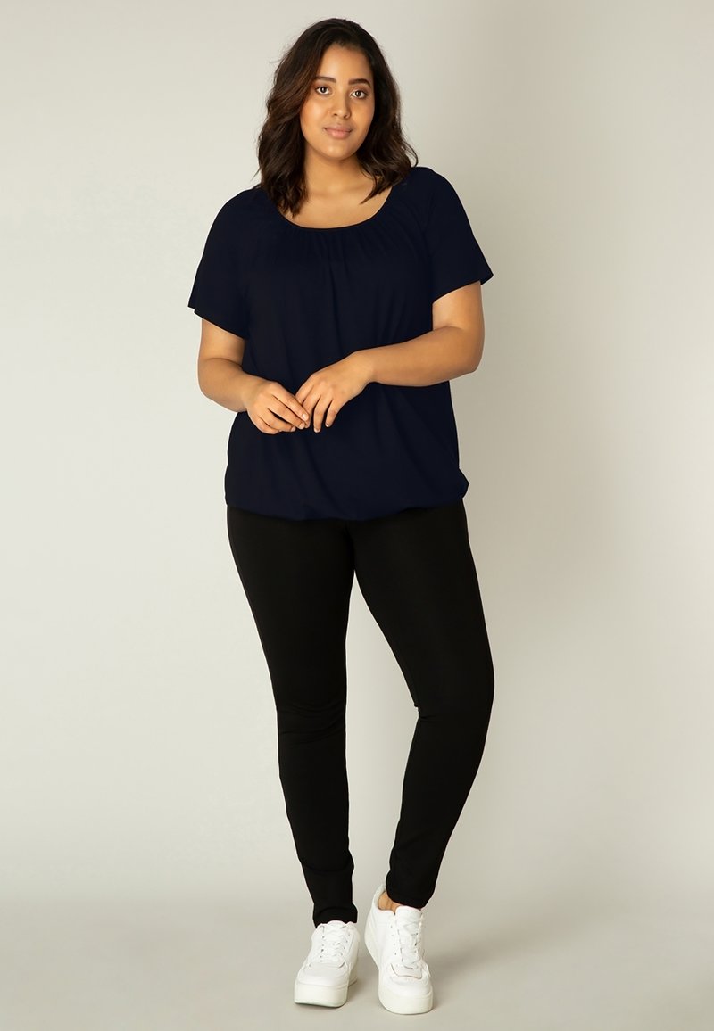 Navyblauw T-shirt met korte mouwen en een ronde hals, gecombineerd met zwarte leggings en witte sneakers. Het shirt heeft subtiele plooien bij de halslijn.