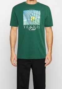 Grön bomullströja med en tennisgrafik som visar en boll och nät, samt texten "Tennis Club" i vitt. Korta ärmar och rundad halsringning.