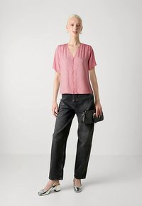 Blouse rose à manches courtes avec un col en V et des boutons à l'avant, associée à un jean noir ample et des chaussures argentées, tenant un sac à main noir.