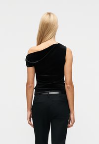 Femme aux longs cheveux blonds portant un haut en velours noir à une épaule et un pantalon noir, debout de dos sur un fond uni.