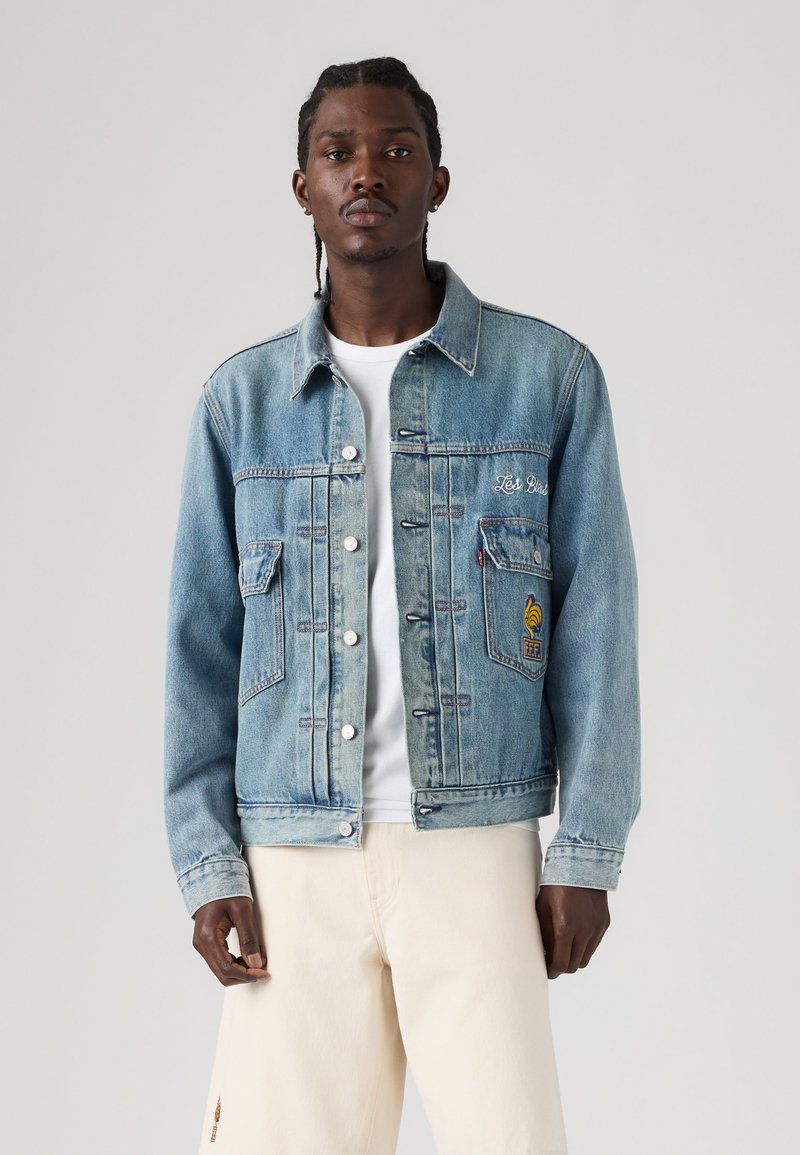 Giovane uomo con capelli intrecciati che indossa una giacca di jeans azzurro chiaro con toppe ricamate, una t-shirt bianca e pantaloni crema, in piedi davanti a uno sfondo grigio.