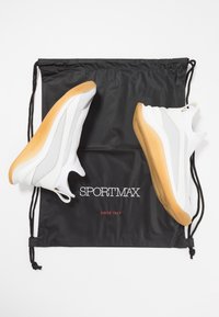Baskets blanches avec des accents gris et une semelle en gomme, posées sur un sac à cordon noir portant le texte "SPORTMAX" en blanc et rouge.