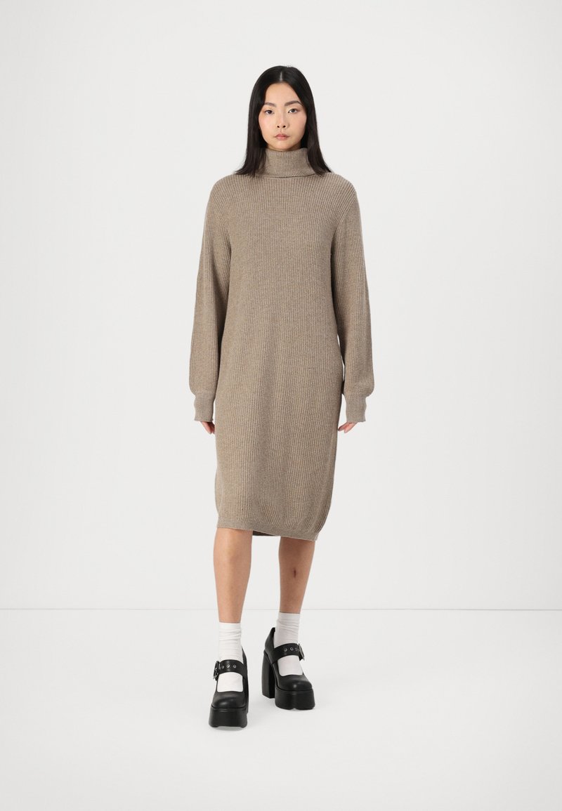 Beige knitted dress with a high collar and long sleeves, featuring a relaxed fit and a textured fabric. Worn with black platform shoes.