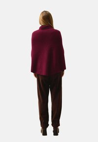 Burgundy stickad poncho med hög krage, ribbad struktur och lös passform, tillsammans med bruna corduroybyxor och svarta klackskor med blockheel.