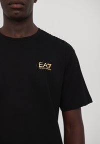 Camiseta de algodón negra con un pequeño logo dorado "EA7 Emporio Armani" en el lado izquierdo del pecho. Diseño de mangas cortas y cuello redondo.