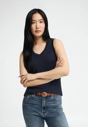 Femme aux longs cheveux noirs portant un haut sans manches côtelé bleu marine et un jean bleu, debout les bras croisés devant un fond uni.