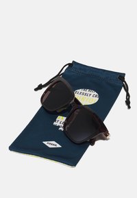 Schildpatt-Sonnenbrille mit quadratischen Rahmen auf einem dunkelblauen Stoffbeutel mit gelbem und weißem Text; der Beutel hat einen Zugverschluss.