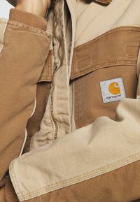 Persoon in een bruine en beige Carhartt-jas met rits, waarbij het merkembleem op de borstzak zichtbaar is.