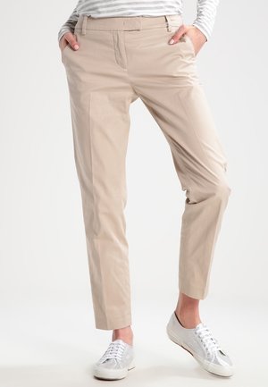 Persoon die beige rechte broek draagt met handen in de zakken, gecombineerd met zilverkleurige sneakers en een grijs-wit gestreept shirt met lange mouwen.