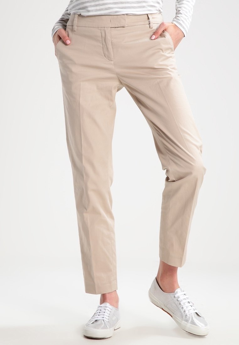 Personne portant un pantalon beige droit avec les mains dans les poches, associé à des baskets argentées et une chemise à manches longues rayée gris et blanc.