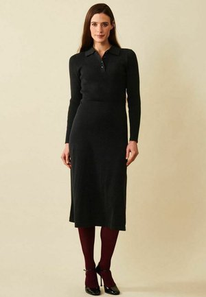 Pitkähiuksinen nainen, jolla on ruskeat hiukset, musta pitkähihaisen midihameen mekko, viininpunaiset sukkahousut ja mustat korkokengät, seisoo beigen taustan edessä.