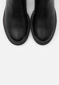 Une paire de bottes en cuir noir avec des orteils arrondis et des coutures visibles le long des bords de la semelle sur un fond clair.