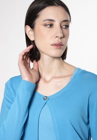 Hellblaue Strickjacke aus weichem Material mit rundem Ausschnitt, langen Ärmeln, einer Knopfverschluss und Spitzenakzenten am Ausschnitt.