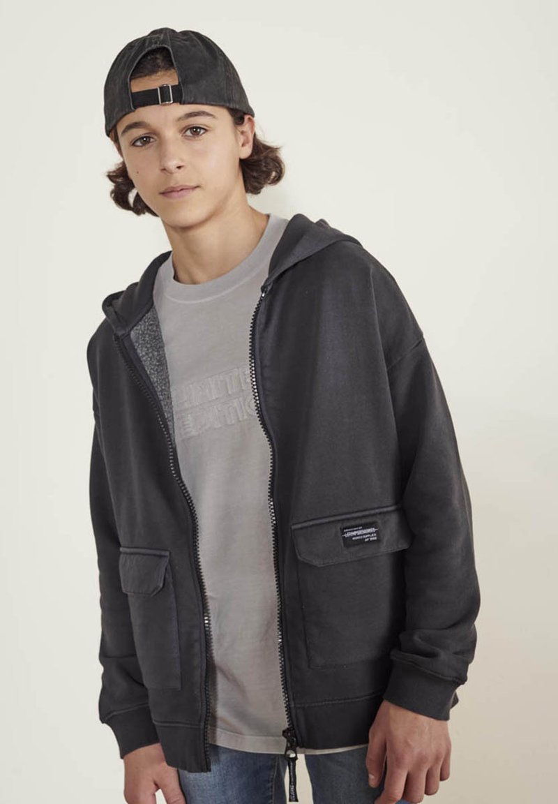 Un sweat à capuche zippé gris foncé avec deux poches avant et une doublure intérieure texturée, porté sur un t-shirt gris clair, avec une casquette noire.