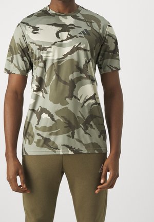 Mann trägt kurzärmliges grünes und braunes Tarn-T-Shirt mit passenden olivgrünen Hosen und steht vor einem schlichten hellen Hintergrund.