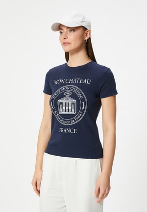 Navyblauwe t-shirt met korte mouwen, met een witte grafische ontwerp en tekst. Gecombineerd met lichtgekleurde broek en een witte pet.