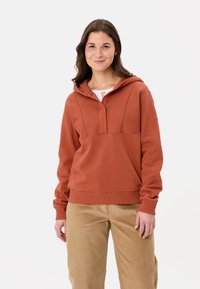 Hoodie couleur rouille avec une poche kangourou, une demi-fermeture éclair et des poignets côtelés. Porté avec un pantalon en velours côtelé beige. Texture lisse et douce.