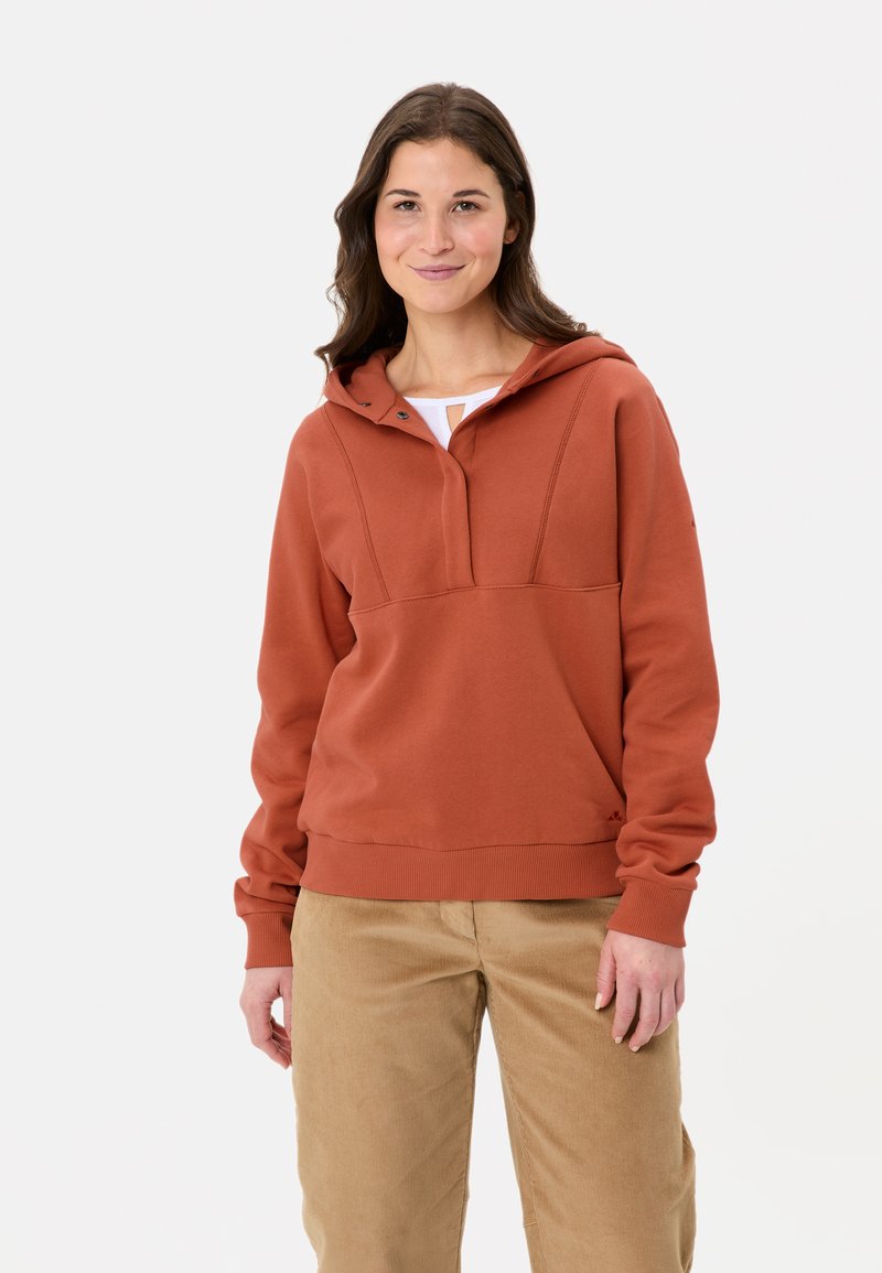 Hoodie couleur rouille avec une poche kangourou, une demi-fermeture éclair et des poignets côtelés. Porté avec un pantalon en velours côtelé beige. Texture lisse et douce.