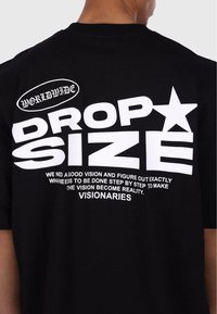 Svart bomulls T-skjorte med fet hvit tekst. Funksjoner inkluderer "DROP SIZE" og grafikk i ulike skriftstørrelser og stiler, inkludert en stjerneakksent.