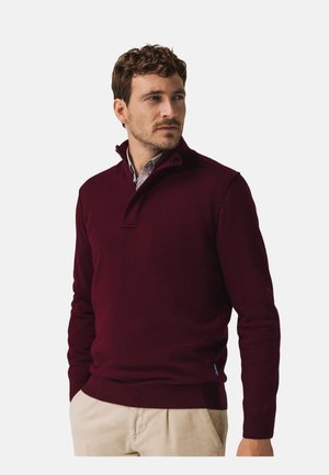 Bordeauxrode gebreide trui met een halflange ritssluiting en twee knopen, gecombineerd met een licht overhemd, gedragen met neutrale ribbroek.