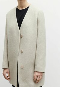 Ljusgrå kappa med texturerat tyg, V-ringning och tre stora beige knappar. Har lång ärm och rak skärning.