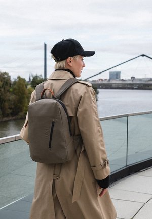 Zaino grigio con dettagli neri e un manico in pelle marrone, indossato sopra un trench beige. Ambientazione urbana vicino all'acqua con un ponte sullo sfondo.