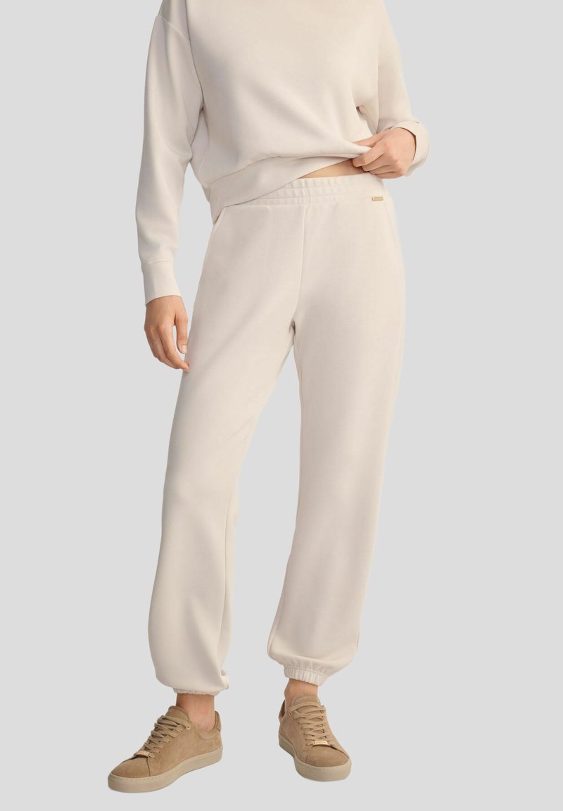 Osoba w kremowych dresach i pasującym swetrze, w zestawieniu z beżowymi sneakersami, stojąca na neutralnym tle.