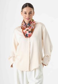 Zijde sjaal in zachtroze met levendige bloemmotieven en een oranje rand, gedragen om de nek met een crèmekleurig button-down shirt.