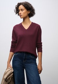 Weinroter Strickpullover mit V-Ausschnitt, lockerer Passform und langen Ärmeln, getragen über dunkelblauen Jeans; hält ein hellbeiges Kleidungsstück.