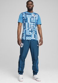 Tricoul sportiv albastru are modele abstracte și un logo, fiind asociat cu pantaloni de culoare albastru închis și ghete de fotbal albe cu accente roz.