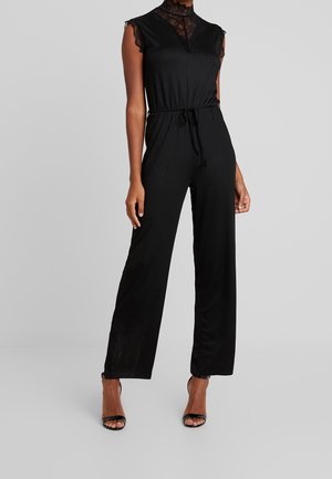 Kobieta w czarnym kombinezonie bez rękawów z koronkowym kołnierzem i wiązanym w pasie, w czarnych sandałkach z odkrytym noskiem, stojąca na jednolitym tle.