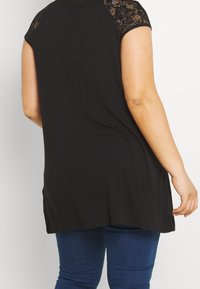 Túnica negra con mangas de encaje, un corte relajado y un dobladillo ligeramente curvado, combinada con jeans de mezclilla oscuros.