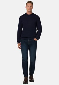 Maglione a maglia color blu scuro con texture a coste, abbinato a jeans denim scuri e stivali in pelle marroni con lacci. Il modello sta in piedi con le mani in tasca.