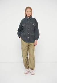 Veste en denim gris avec deux poches poitrine, boutons à pression et coutures contrastantes ; portée avec un pantalon cargo beige clair et des baskets roses.