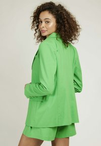 Blazer vert en tissu léger, doté d'une poche unique, d'un col cranté et de poignets boutonnés. Associé à un short assorti.