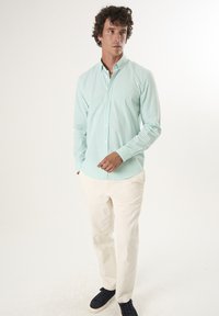 Camisa de algodón a cuadros de color verde claro, con mangas largas, cuello abotonado y dobladillo curvo, combinada con pantalones de color crema y zapatillas oscuras.