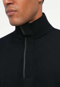Schwarzer Strickpullover mit hohem Kragen und halbem Reißverschluss an der Vorderseite. Mit gerippter Struktur und schmaler Passform, die ein modernes Design betont.