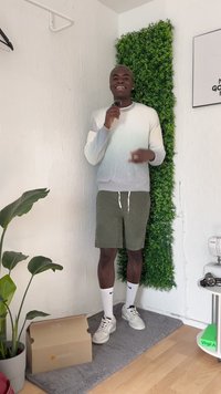Maglione con gradiente da grigio chiaro a bianco, pantaloni corti color oliva, calze bianche e scarpe da ginnastica. Pianta in vaso e scatola di cartone in una stanza con parete verde.
