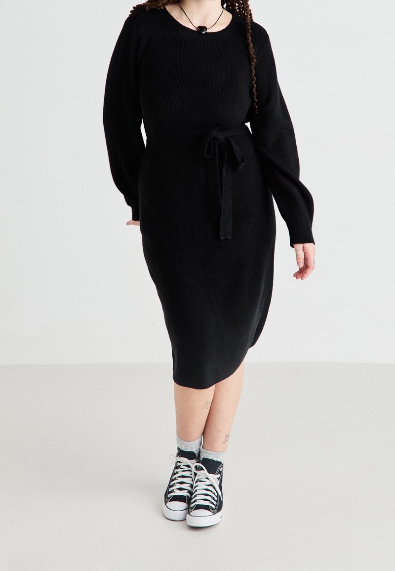 Femme portant une robe en tricot noire avec une taille nouée et des baskets hautes noires, se tenant devant un fond blanc.