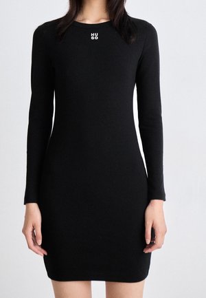 Kobieta w dopasowanej czarnej sukience z długim rękawem, z logo "HUGO" umieszczonym na środku dekoltu, na tle jednolitego jasnego tła.