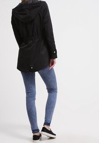 Black jacket with hood, cinched waist, and gold-tone button accents, paired with blue denim-like skinny jeans and black slip-on shoes.