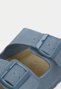 Κοντινό πλάνο από ιμάντες μπλε σουέτ σανδαλιών με ρυθμιζόμενες μεταλλικές αγκράφες και ορατό ταμπά πάτο από κάτω.