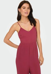 Combinaison rouge-bourgogne avec des bretelles fines et un décolleté en V. Elle présente une coupe décontractée et des poches latérales pour plus de fonctionnalité.