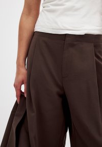 Pantalones tailoring marrones hechos de una tela suave. Incluyen características como un frente plano, bolsillos laterales y un corte holgado que fluye desde las caderas.