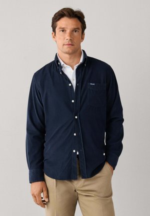 Hombre con cabello castaño que lleva una camisa azul marino abotonada sobre una camisa blanca y pantalones beige, de pie frente a un fondo claro y liso.