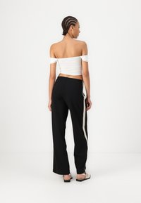 Haut blanc à épaules dénudées en tissu texturé, associé à un pantalon noir à jambes larges avec une bande latérale beige. Des chaussures plates noires complètent le look.