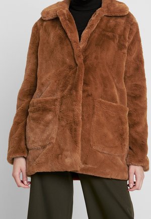 Manteau en fausse fourrure marron avec un large col et deux grandes poches avant, porté sur un top noir et un pantalon foncé.