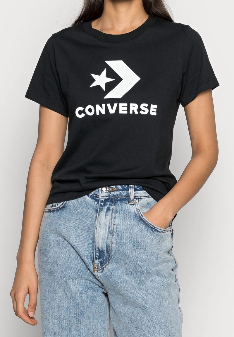 Femme portant un t-shirt Converse noir avec logo blanc et un jean taille haute bleu clair, une main dans la poche, sur fond blanc.
