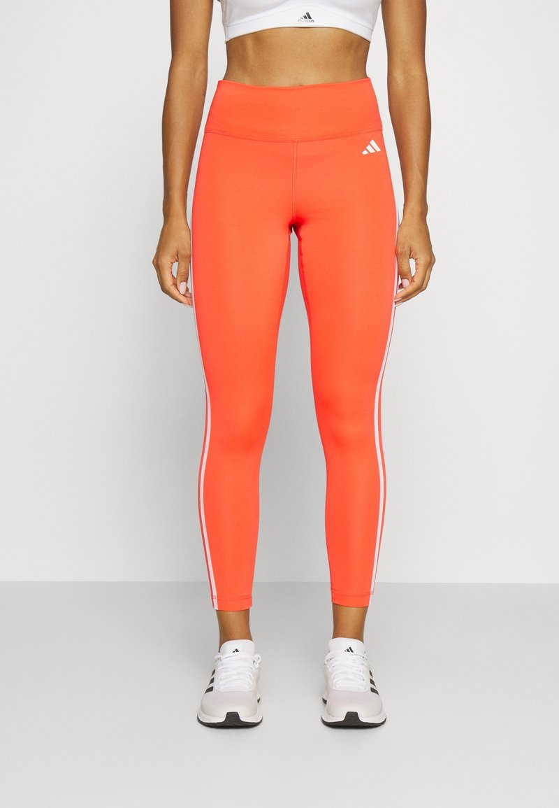 Leggings taille haute, ajustés, de couleur orange vif avec des bandes latérales blanches et un tissu lisse et extensible. Associés à des chaussures de sport blanches.